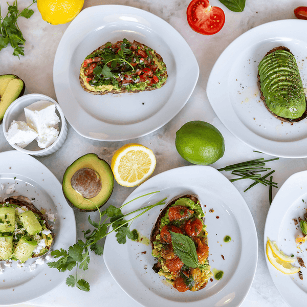 Avocado Toast Top Shot: A variety of Avocado toasts plated on white serving plates surrounded by key ingredients on a kitchen countertop