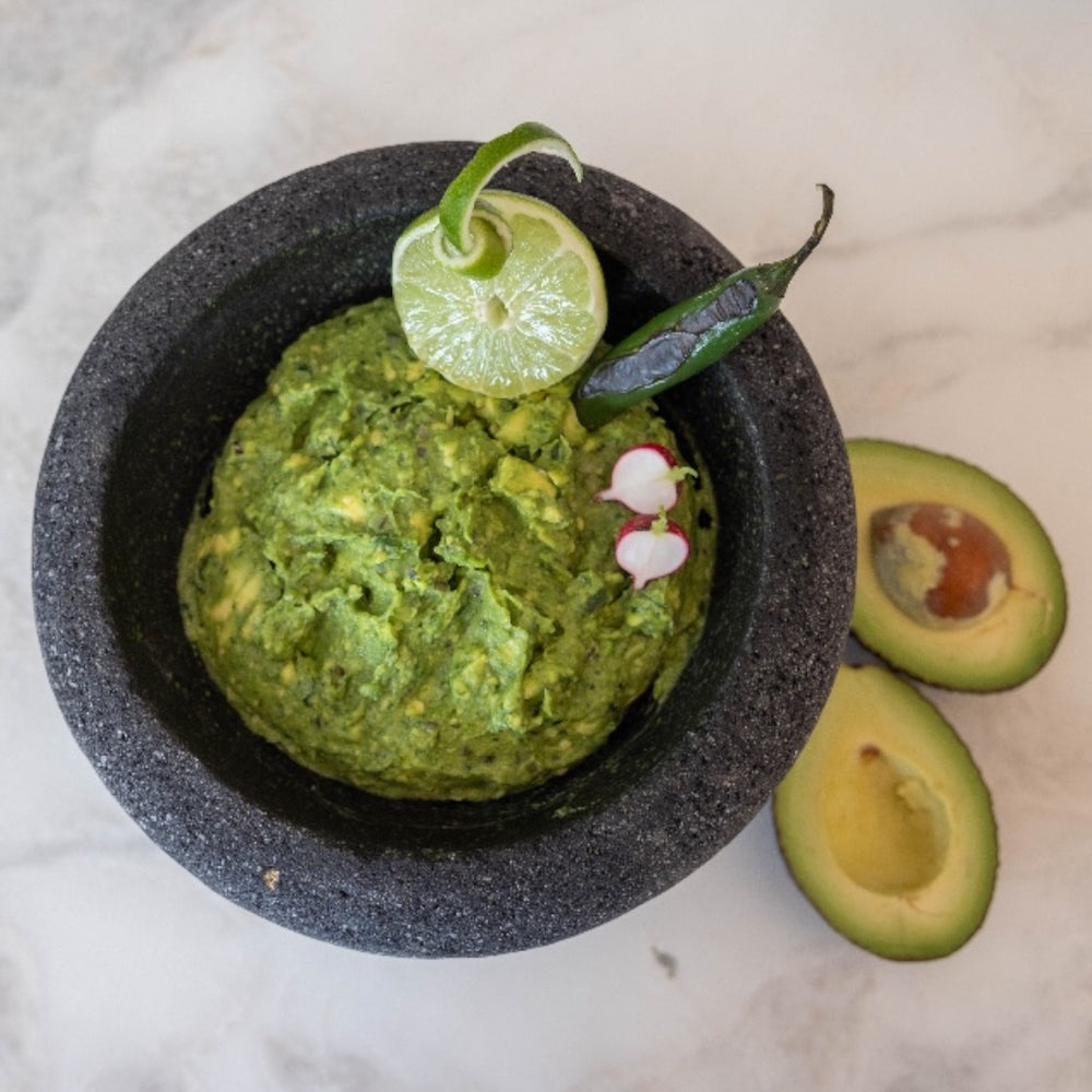 Guacamole served in lava rock molcajete with open avocado displayed to its side.