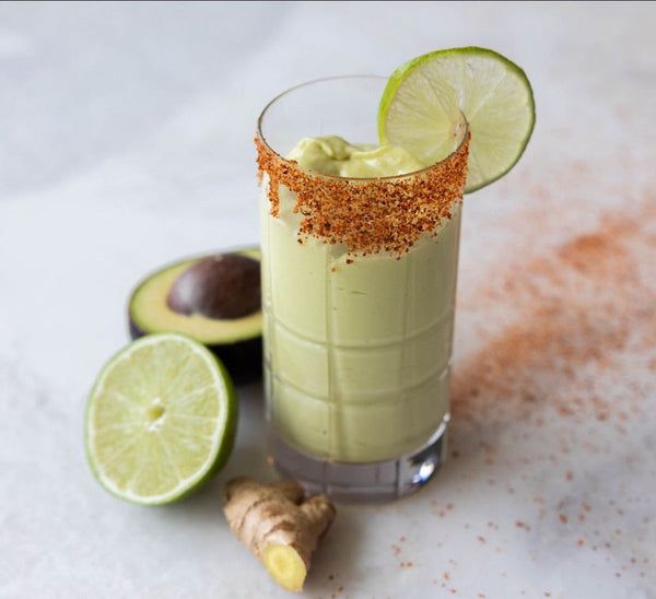 a creamy avocado smoothie garnished with a lime slice and chili powder rim, surrounded by fresh lime and ginger on a light background