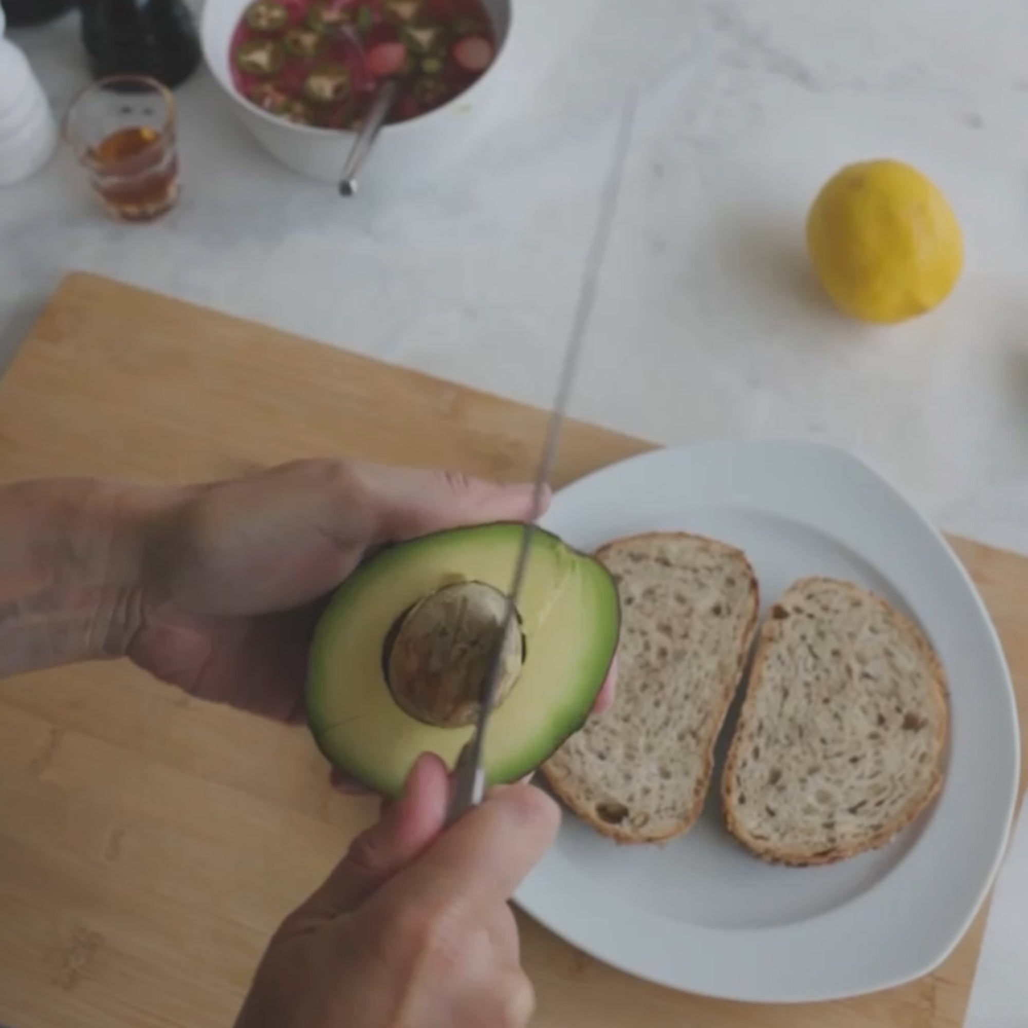 a person slicing an avocado on a plate with two slices of bread, while an orange is seen in the background
