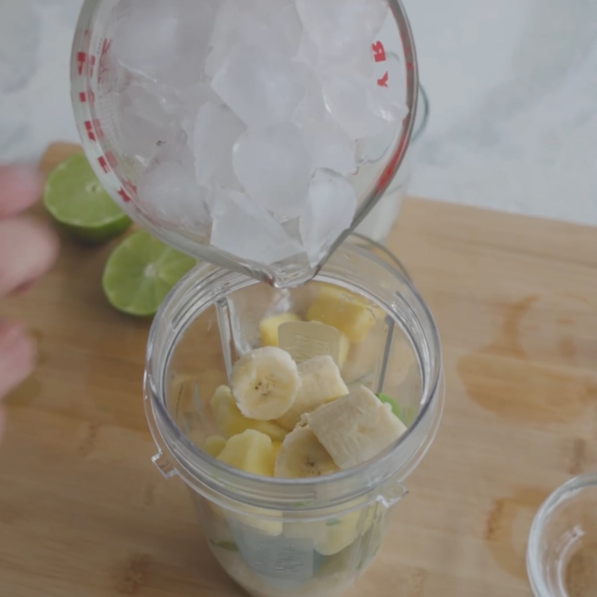 ice being pourred into a blender containing banana slices, with lime halves visible in the background