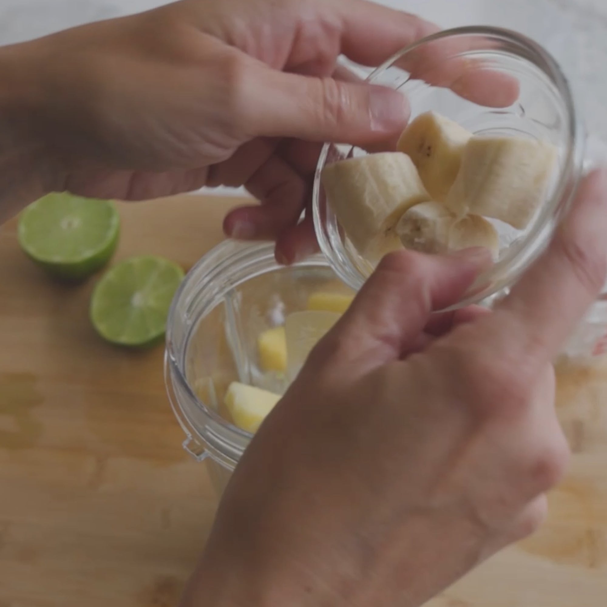 a person adding banana slices from a small bowl into a blender, with lime halves visible on the side
