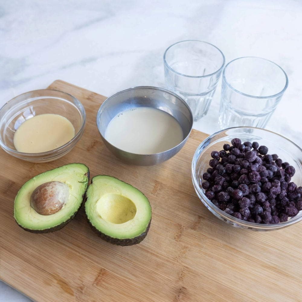 ingredients on a cutting board: halved avocado, blueberries, and bowls of creamy components
