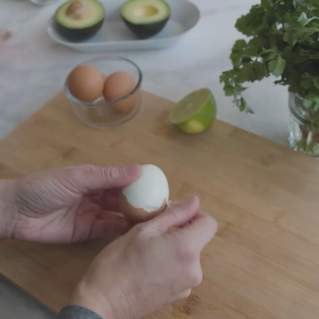 peeling a hard-boiled egg on a cutting board, with avocado and eggs in the background