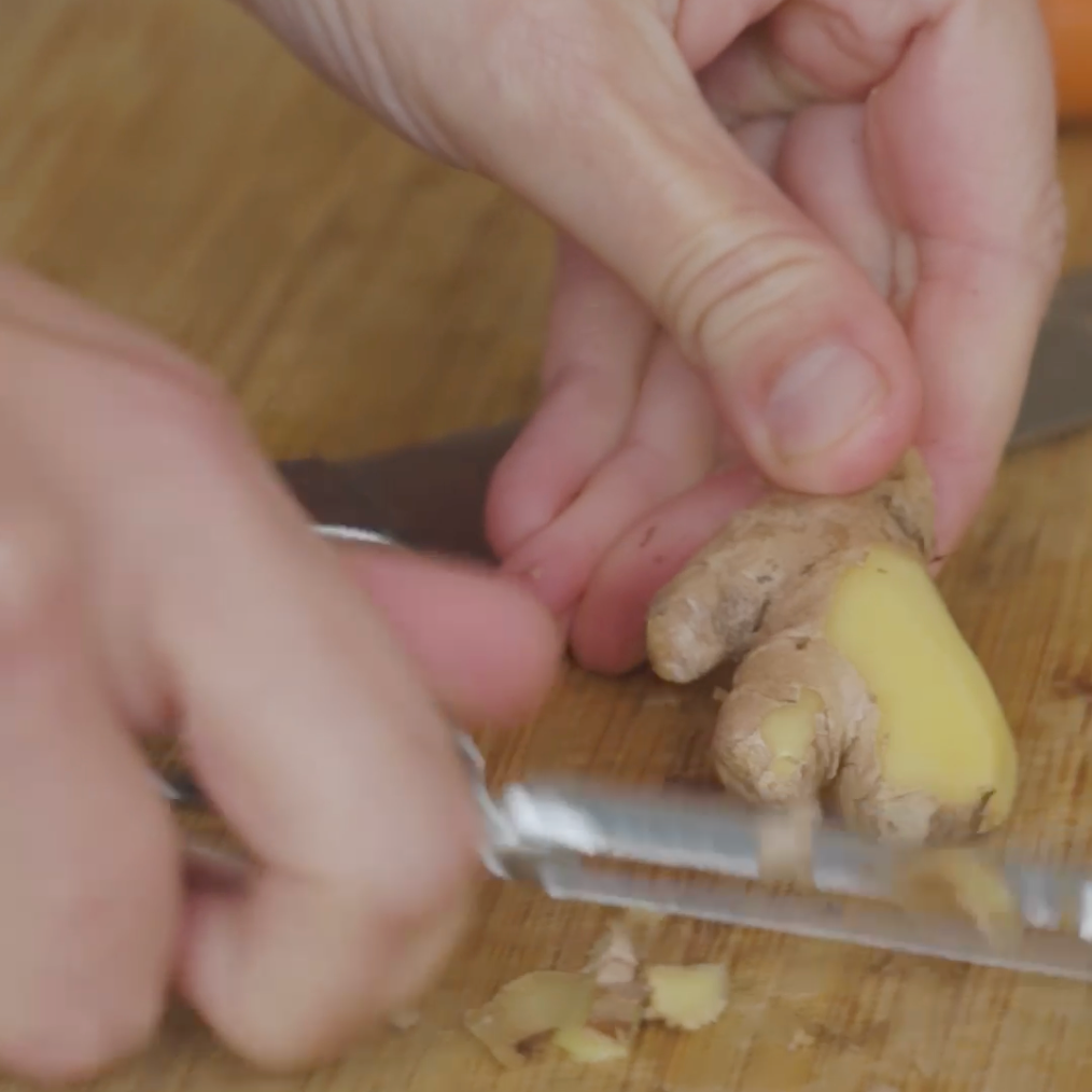a person peeling ginger root with a knive on a wooden cutting board
