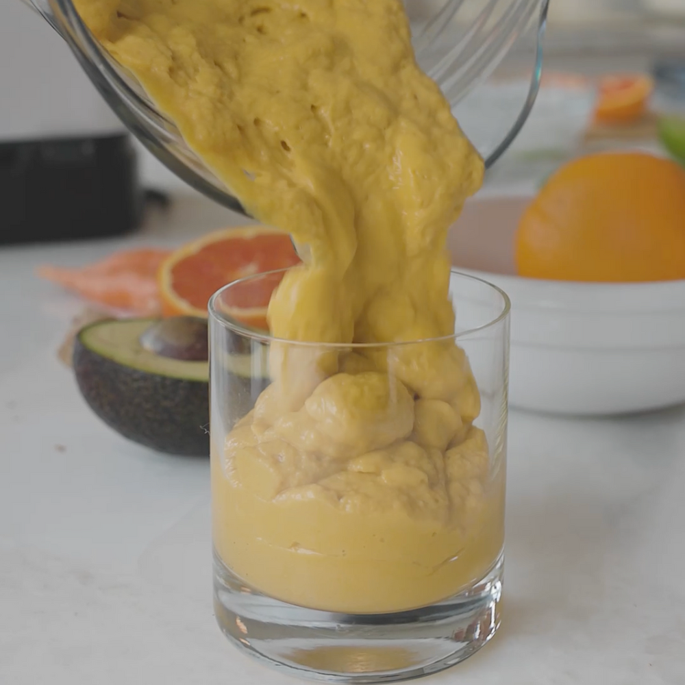a thick smoothie or juce being poured from a blender into a glass, with orange slices in the background