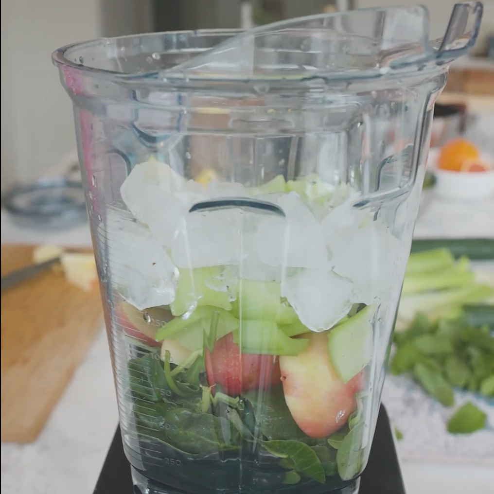 a blender filled with ingredients like apple slices, avocado, and spinach leaves, ready for blending with ice on top