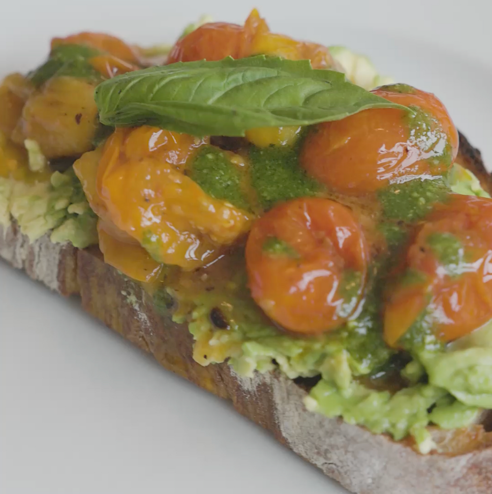 a slice of bread topped with mashed avocado, cherry tomatoes, pesto, and a basil leaf