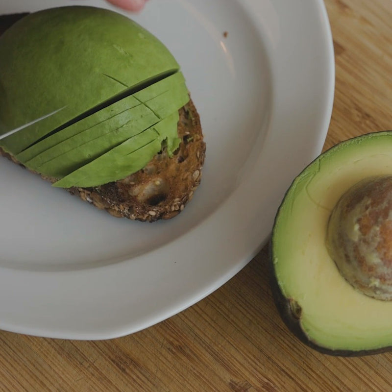 an avocado being sliced on a white plate, with one half showing the seed and the other half partially sliced