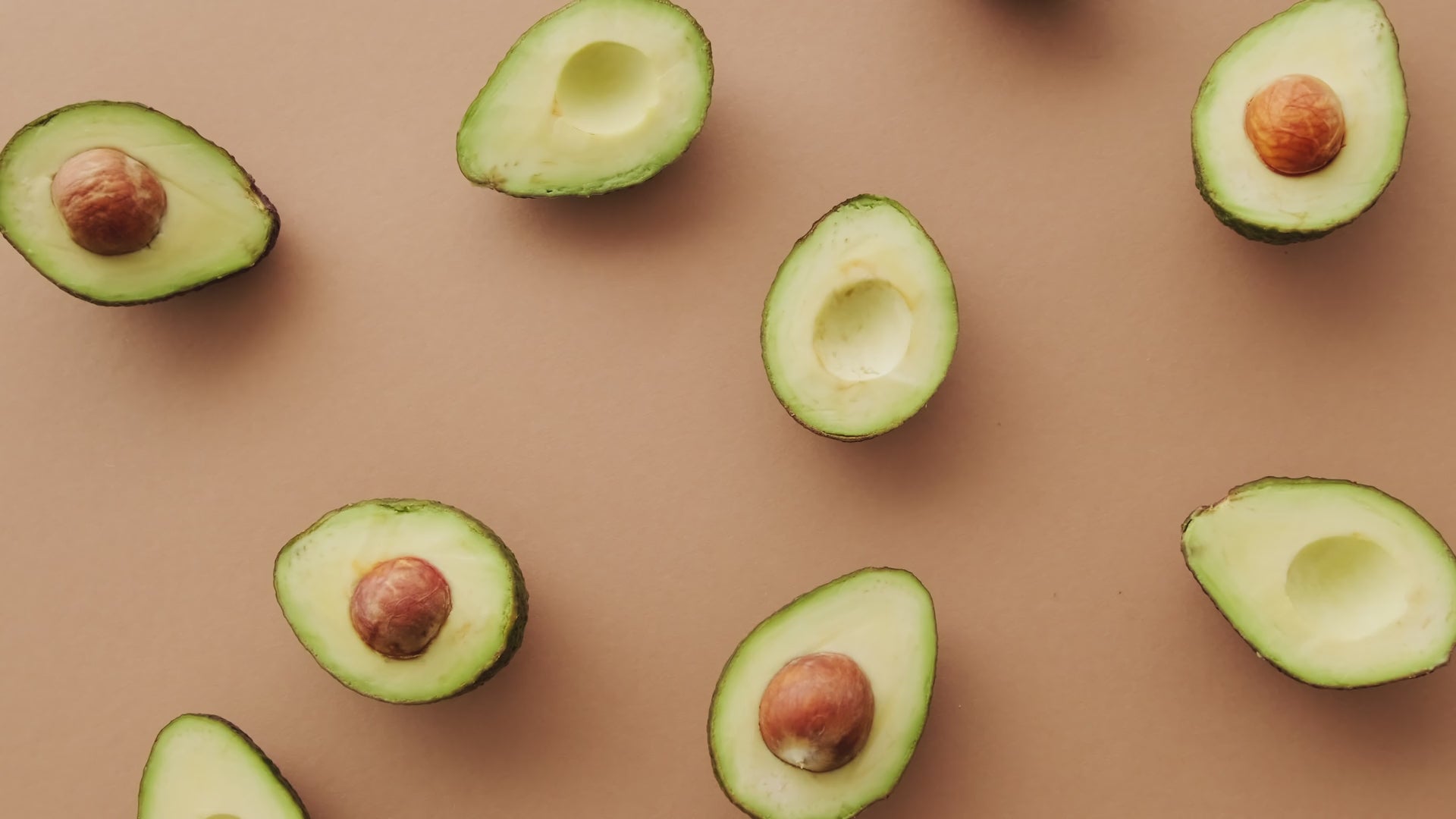 Top-down video of numerous halved avocados arranged in a circular pattern. The camera slowly rotates, capturing the creamy green flesh and brown pits against a salmon-pink background.